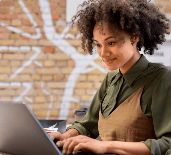 Mulher negra de camisa verde e jardineira marrom digitando no teclado do notebook.