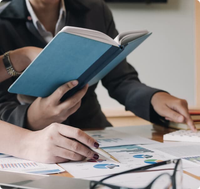 Duas mãos femininas, sendo que uma aponta para um caderno aberto na mão de um homem e a outra segura uma caneta sobre um papel; e duas mãos masculinas, sendo que uma segura um caderno na mão e a outra tecla em um teclado.