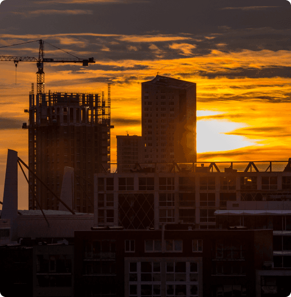 Prédios e guindastes dando a entender que é um pátio de obras, com um céu de entardecer em uma cidade.