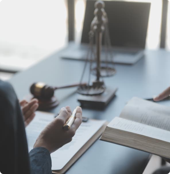 Mãos masculinas de dois homens em um escritório de advocacia, sendo que uma das mãos de um deles segura uma caneta e as mãos do outro homem segura um livro, enquanto há papéis, uma balança do Direito, um martelo e um notebook sobre uma mesa.