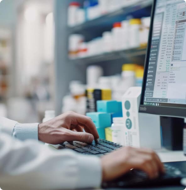 Mãos masculinas digitam em um teclado de computador com parte do monitor à mostra e estantes brancas ao fundo repletas de medicamentos.