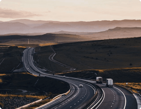 Uma rodovia com faixas estreitas e mata baixa dos lados com poucos automóveis passando, com um céu parcialmente nublado.