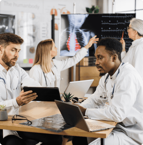 Um homem branco segura um tablet apontando para alguma informação na tela enquanto um homem negro observa a mesma tela, ambos estão sentados um de frente para o outro com uma mesa entre eles. Ao fundo, duas mulheres brancas conversam, enquanto uma está em pé apontando uma caneta para um quadro com gráficos médicos e a outra aponta um dedo para o mesmo quadro. O cenário é uma sala de clínica ou hospital.