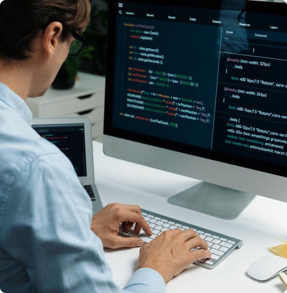 Homem branco de óculos e camisa branca digita em um teclado de um iMac com um notebook ao lado do monitor.