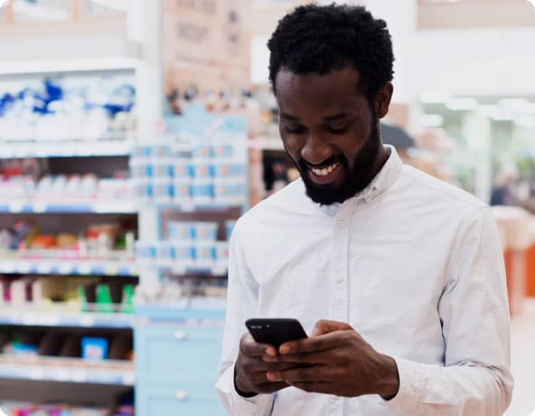 Homem negro com camisa branca segurando um celular com as duas mãos enquanto sorri, com prateleiras repletas de itens à venda ao fundo que remetem a um supermercado.