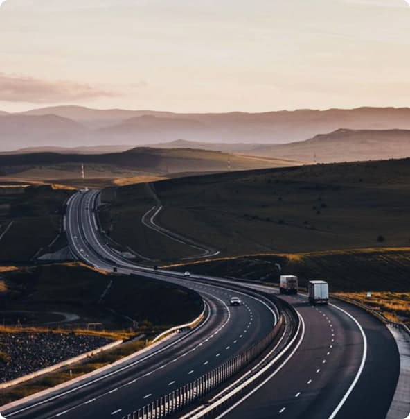 Uma rodovia com faixas estreitas e mata baixa dos lados com poucos automóveis passando, com um céu parcialmente nublado.