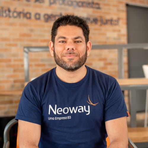 Michel Ávila, CDO da Neoway, homem branco com cabelo e barba escuros, camiseta azul com o logo da Neoway, olhando para frente de maneira sorridente.