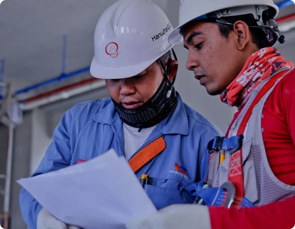 Dois homens de capacete e uniformes de construição civil seguram um papel.