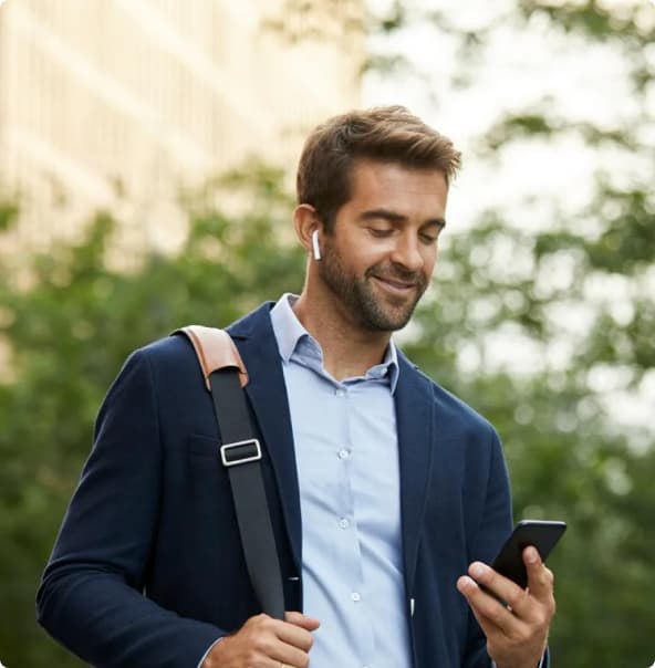 Homem branco vestindo camisa azul clara e terno azul marinho em pé enquanto segura um celular com uma das mãos, enquanto segura a alça de sua mochila com a outra e está ouvindo algum áudio com os fones de ouvido.