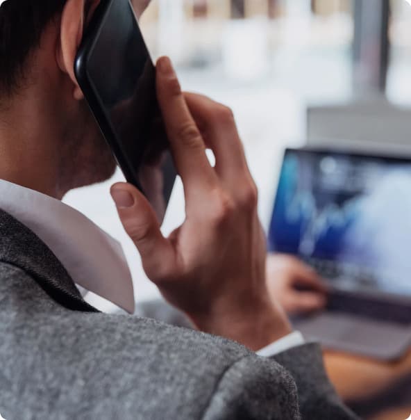 Um homem branco segura um celular com uma das mãos e a outra está digitando no teclado do notebook.