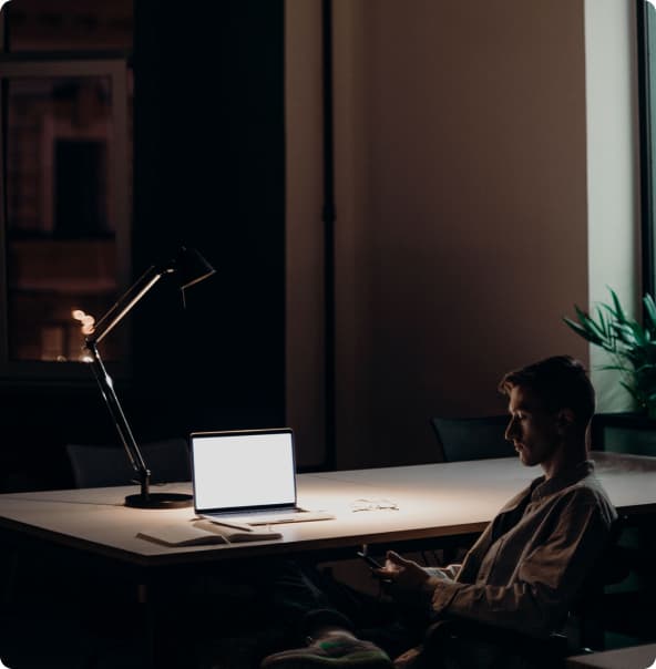 Um homem branco sentado segurando um celular em uma sala com luz baixa, com uma mesa próxima ao seu corpo, a qual possui uma luminária, uns cadernos e um notebook sobre ela, com uma planta perto da parede.