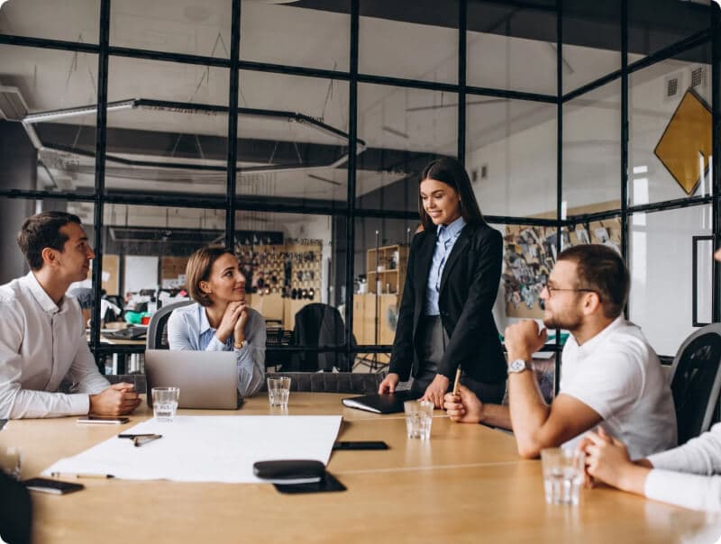 Uma mulher em pé em uma sala de reuniões conversa com um grupo de pessoas, o qual é composto por outras duas mulheres e dois homens, todos sentados e com os braços apoiados em uma mesa de escritório que possui copos, papéis, canetas e outros objetos.