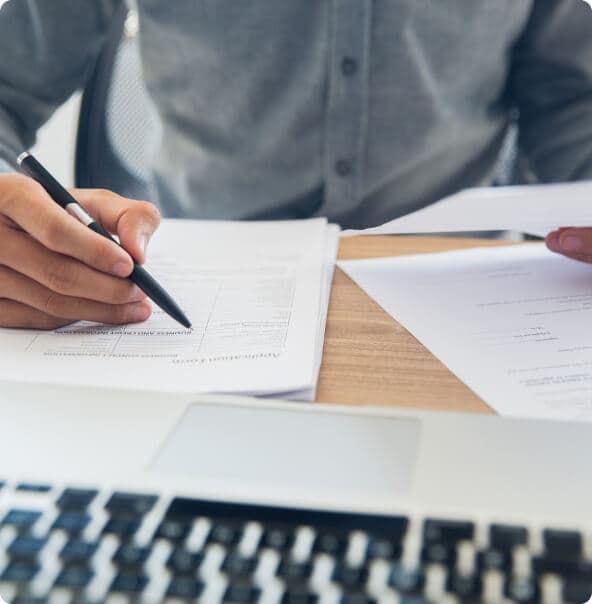 Mão de um homem segura uma caneta enquanto escreve em um papel e a outra mão segura um outro papel, os quais estão apoiados sobre uma mesa com um notebook.