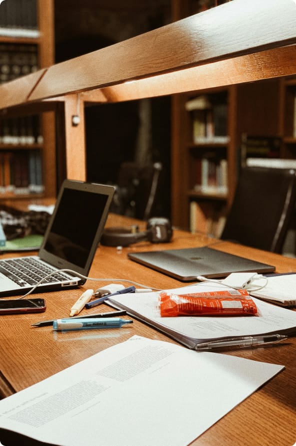 Uma mesa que aparenta ser um escritório de advocacia com vários papéis, canetas e notebooks espalhados sobre ela, com estantes com livros ao fundo.