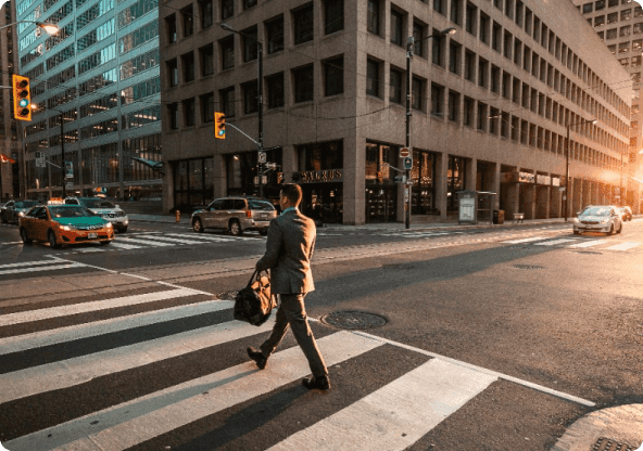 Um homem segurando uma mala e vestindo um terno atravessa a rua na faixa de pedestres em uma cidade com prédios ao fundo e carros parados esperando o semáforo abrir.