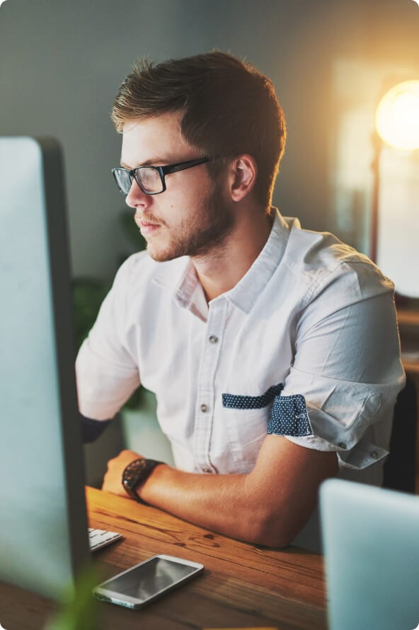 Um homem branco de óculos está olhando para o monitor de seu computador com um braço sobre a mesa. Essa mesma mesa está com o teclado do computador e um celular sobre ela.