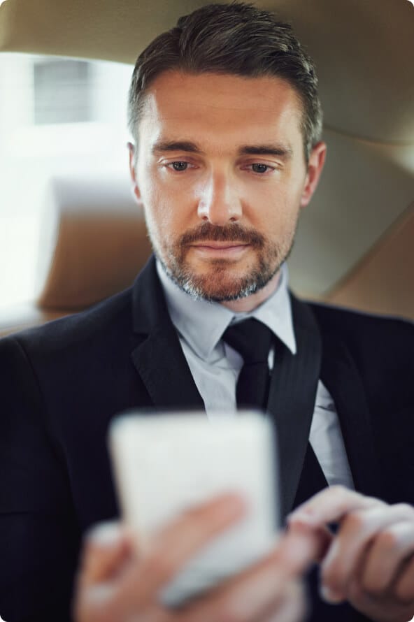 Um homem branco com barba e terno segura um celular com uma das mãos e a outra está posicionada para teclar na tela do aparelho.