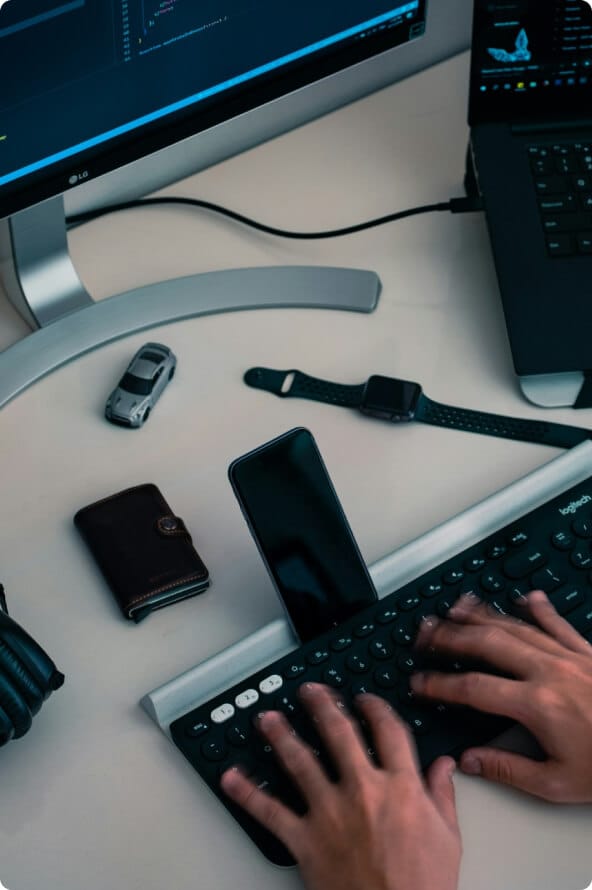 Mãos de uma pessoa digitam em um teclado de computador com um celular em um apoio em sua frente. O teclado está sobre uma mesa com carteira, carrinho de brinquedo, relógio, monitor e notebook.