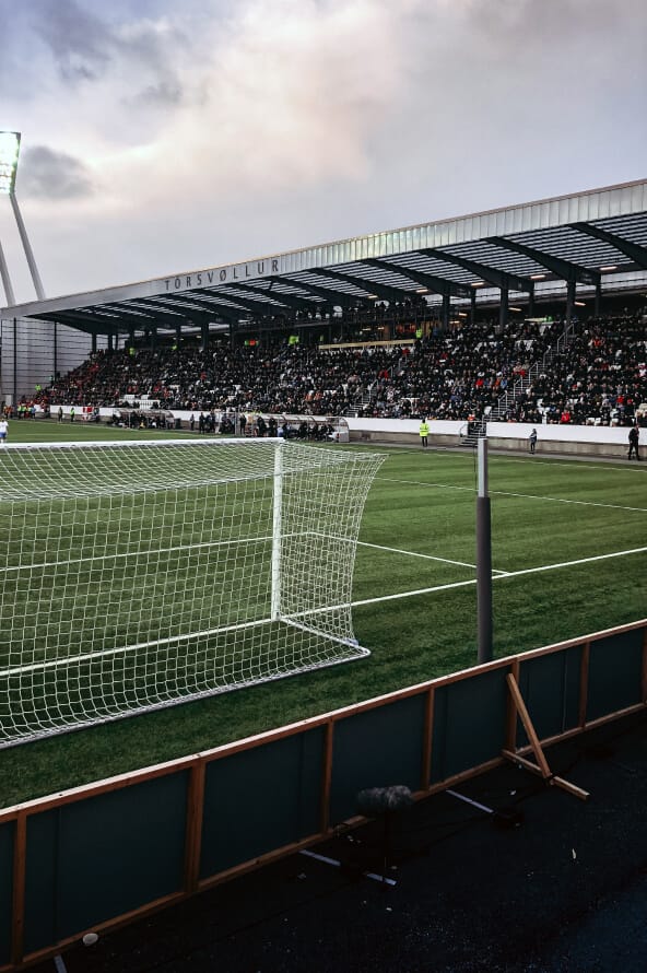 Campo de futebol lotado de pessoas em uma das arquibancadas da direita do campo, com uma das traves e céu encoberto por nuvens.