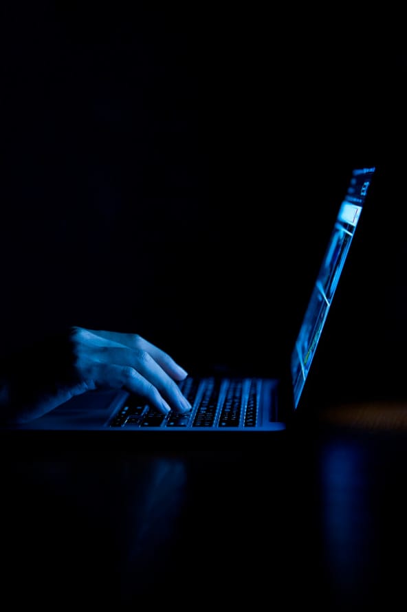 Mãos de uma pessoa digita em um teclado de notebook em um ambiente escuro em tons de preto e azul escuro.