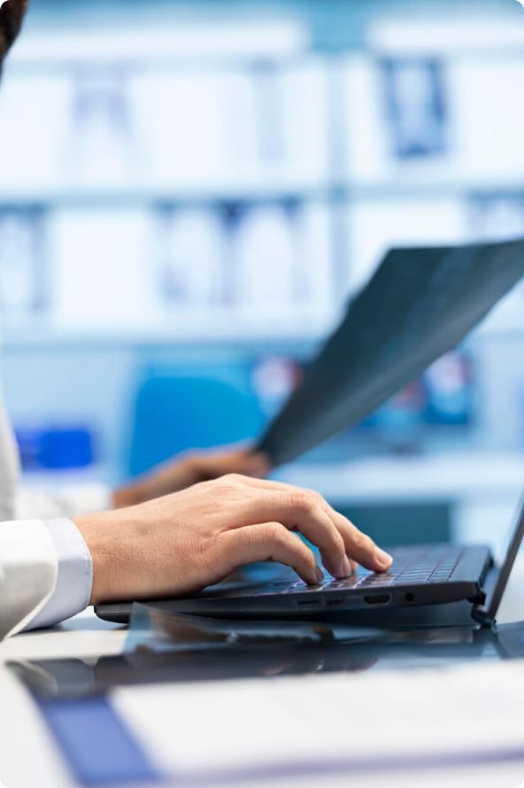 Uma mão de um homem segura um papel enquanto a outra digita no teclado de um notebook.