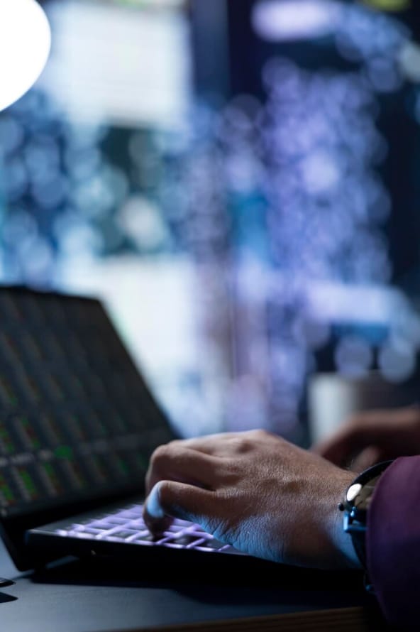 Mãos de uma pessoa digitam em um teclado de notebook em um ambiente com luzes indecifráveis ao fundo.